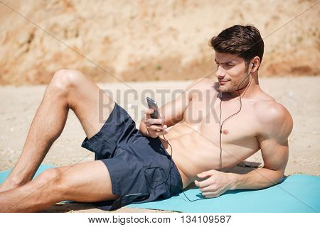 Attractive athletic young man lying and listening to music from mobile phone on the beach