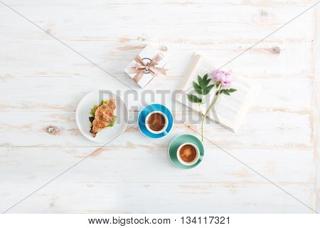 Top view of two cups of coffee, croissant, present box, book and flower on wooden table