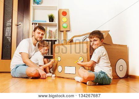 Two kid boys with their father painting headlights on toy cardboard car with yellow felt-tip pens at the room