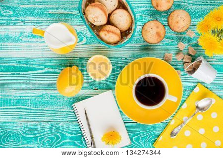 cup of black tea on yellow plate and yellow milk jug cane sugar notebook pen cakes dandelions teaspoon on turquoise colored wooden table with yellow napkin at polka dots top view