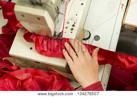 Tailor's hand on sewing machine fastening red satin ribbon on cloth.