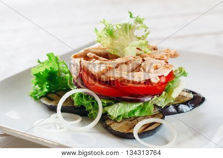 Layered salad with chicken, tomato and grilled eggplant on white. White square plate with portion of layered salad with fried chicken strips, tomato, lettuce, onion and grilled eggplant.