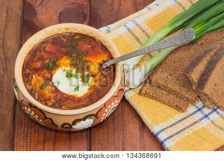 Ukrainian borscht garnished with dill and a sour cream in ceramic bowl and a spoon in it on the background of a green onions and sliced brown bread on a chequered linen cloth on wood planks closeup