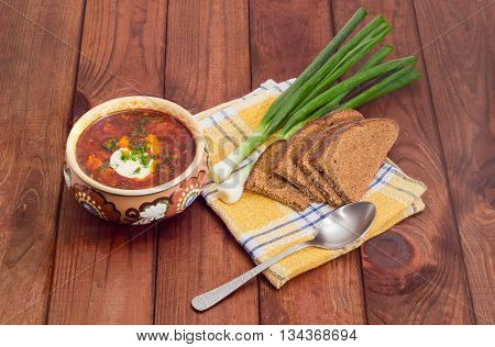 Ceramic bowl of Ukrainian borscht garnished with dill and a sour cream green onions sliced brown bread and spoon on a chequered linen cloth on wood planks