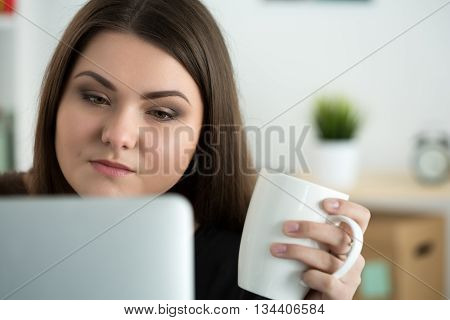Portrait of young sad or attentive woman looking at laptop monitor and holding white cap of tea. Online education coffee break or dieting concept