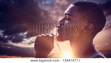 Profile portrait of sportsman is kissing his medal against blue and orange sky with clouds