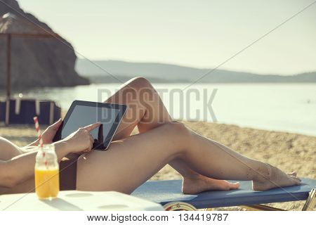 Woman lying on a sunbed on the beach drinking orange juice and surfing the web on a tablet computer while sunbathing in the morning sun. Focus on tablet