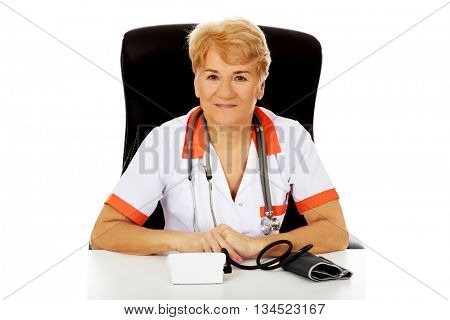 Smile elderly female doctor or nurse sitting behind the desk with bloog preasure gauge