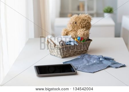 babyhood, motherhood, clothing, technology and object concept - close up of baby clothes and toys for newborn boy in basket with tablet pc computer at home
