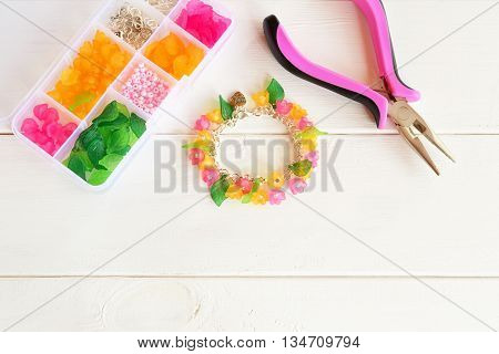 Bright beautiful hand bracelet with pink and yellow acrylic beads flowers and green leaves. Pliers. Organizer with beads, plastic flowers and metal elements for jewelry on white wooden background