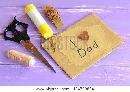 Hand greeting card with message I love dad. Homemade design fathers day card. Happy birthday card for dad. Scissors, glue, cord, thread, needle. Craft inspirations for kids. Lilac wooden background