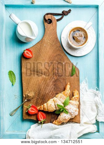 Breakfast or dessert set. Freshly baked croissants with strawberries, cup of coffee and milk in creamer on brown wooden board over blue background, top view, copy space