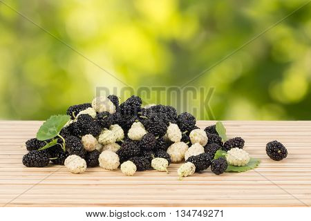 Closeup heap of ripe white and black mulberries with green leaves on table on green leaves background. Horizontal