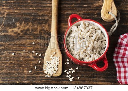 The red ceramic saucepan with white polka dots complete crumbly barley porridge on a simple wooden background with a red cloth in a white cage with a wooden spoon full of dry grain of pearl barley. selective focus
