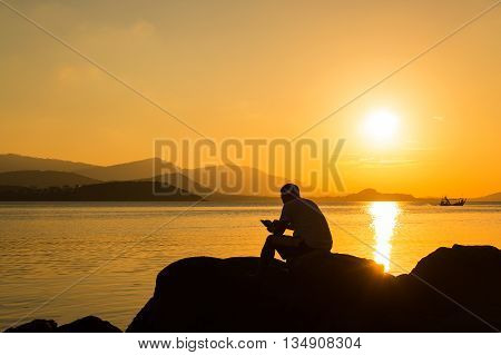 Young man sitting on the stone play smartphone at sea sunset background