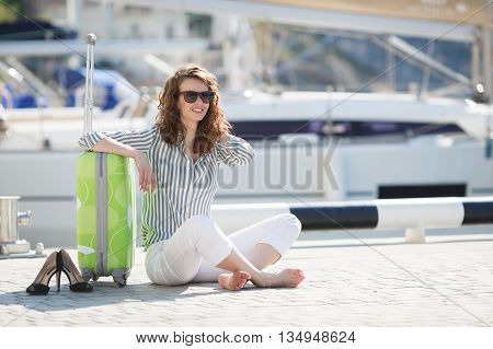 Cute young woman with gray eyes, curly red hair and a sweet smile, dressed in white trousers and a light shirt with stripes, sitting alone on the pier near the yacht club, with a light-green suitcase with wheels on a background of yachts and boats near th