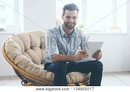 Man with digital tablet. Confident young handsome man holding digital tablet and smiling while sitting in big comfortable chair at home