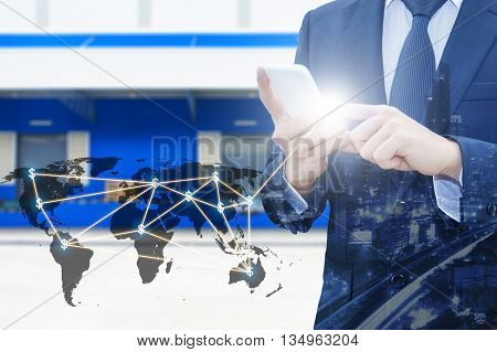 Double exposure of businessman using smart phone network connection world map and city of business center with blurred cargo distribution warehouse background international transportation trading business concept