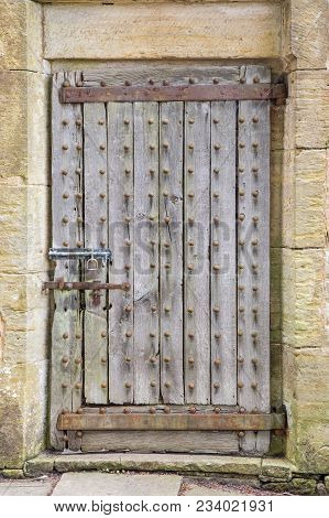 Aged Strong Wooden Door With Iron Studs And Rusty Hinges, Bolted Closed. Framed By Stone Wall.