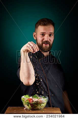 Portrait Of A Handsome Mischevious Male Chef Cook In Black Uniform Making Delicious Vegetable Salad 