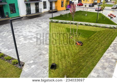 Lawn Mower Cutting Grass On Green Field In Yard Near Apartment Residential Building. Mowing Gardener