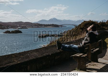Man Sitting On A Bench, Relaxing In The Sun In Kyle Of Lochalsh, Scotland. 
Skye Bridge And Isle Of 