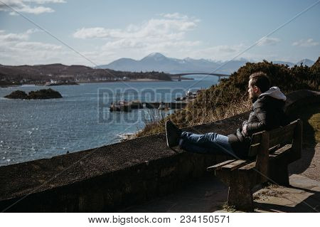 Man Sitting On A Bench, Relaxing In The Sun In Kyle Of Lochalsh, Scotland. 
Skye Bridge And Isle Of 