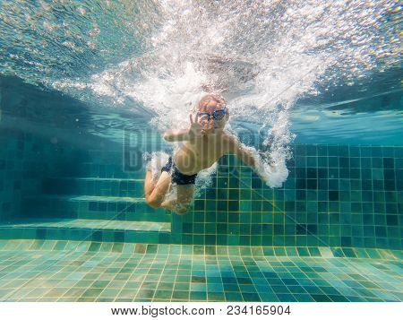 A Child Boy Is Swimming Underwater In A Pool, Smiling And Holding Breath, With Swimming Glasses.