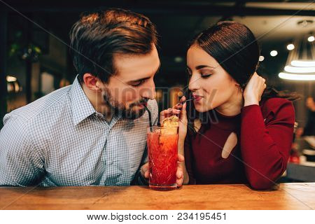 Nice Picture Of Young Couple Sitting Together And Drinking Red Cocktail From The Same Glass At The S