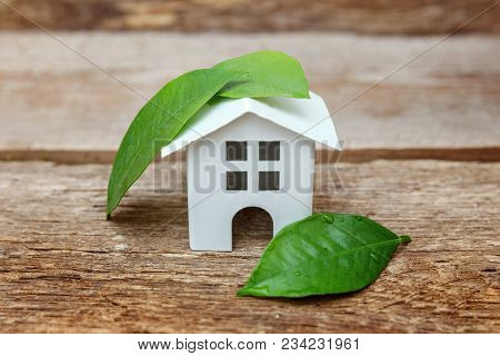 Miniature White Toy Model House With Green Leaves On Wooden Backgdrop. Eco Village, Abstract Environ