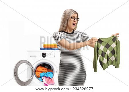 Shocked young woman holding a shrunken blouse in front of washing machine isolated on white background