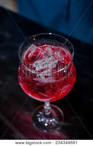 The Bartender Makes A Red Cocktail. Blue Dark Background. A Glass With Ice And Red Drink. Contrast P