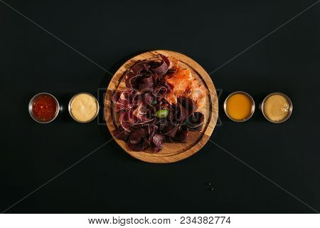 Top View Of Delicious Sliced Assorted Meat On Wooden Board With Various Sauces On Black