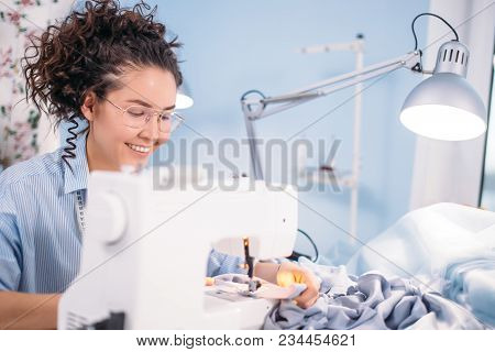 Good Looking Seamstess With Wavy Hair And Glasses Showing Master-class On Sewing Indoors. Teach To C