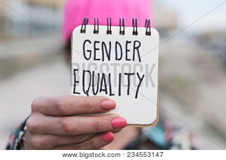 closeup of a young woman outdoors wearing a pink pussycat hat showing a notepad in front of her face with the text gender equality written in it