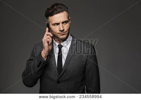 Photo of masculine man 30s dressed in businesslike costume working in office and talking on phone about business isolated over gray background