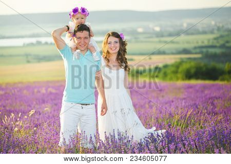 Mom, Dad And Their Little Daughter In A Beautiful Wreath Are Walking In A Lavender Field. Life In Pr