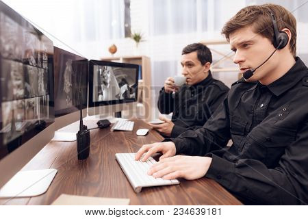 Two Men Work As Guards. They Sit In Front Of The Monitors In The Security Room. The Headphones Are O