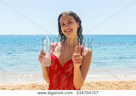 Happy Tanned Girl Holding A Water Bottle Gesturing Thumbs Up On The Beach.
