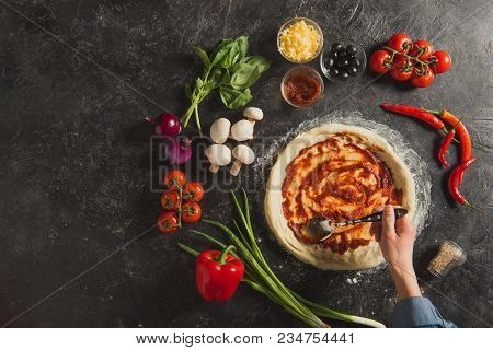 Cropped Shot Of Woman Putting Sauce On Raw Dough While Cooking Italian Pizza On Dark Tabletop