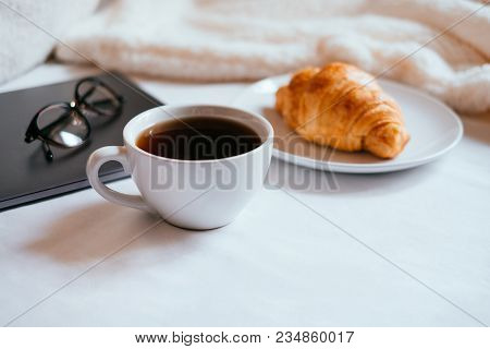 Big White Morning Cup Of Coffee And Crossant In Bed With Notebook, Glasses, Pillow And Cosy Blanket.