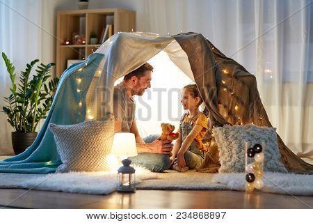 family, hygge and people concept - happy father with teddy bear toy and little daughter playing in kids tent at night at home