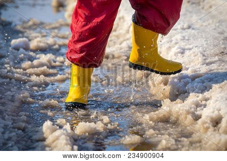 Kid Legs In Yellow Rainboots Running In The Ice Puddle With Melting Snow At Sunny Spring Day, Outdoo