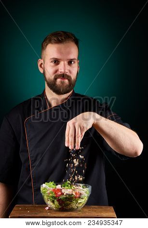 Portrait Of A Handsome Mischevious Male Chef Cook In Black Uniform Making Delicious Vegetable Salad 