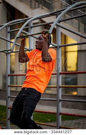 African-american Man In Orange T-hirt In Wireless Headphones Pulling Himself Up On Horizontal Bars O