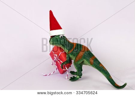 Funny Green Dinosaur Toy In Little Santa Claus Hat  Holding Red Christmas Gift Box