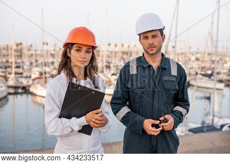 Professional Young Male And Female Marine Colleagues In Hardhats And Workwear With Clipboard And Vhf