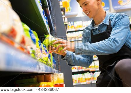 Store Worker Man Smiling And Sitting On One Knee Down Putting Products On Shelves At Supermarket Hol