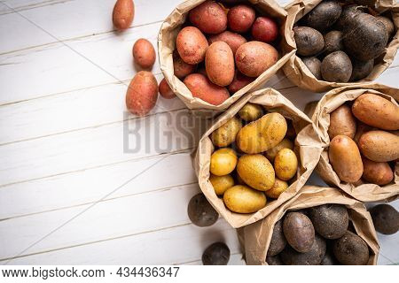 Various Varieties Of New Raw Colorful, White, Red And Purple Potatoes In Paper Bags On White Wooden 