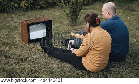 Family Of A Man, A Woman And Little Baby Are Sitting On The Grass And Watching An Old Retro Tv. The 